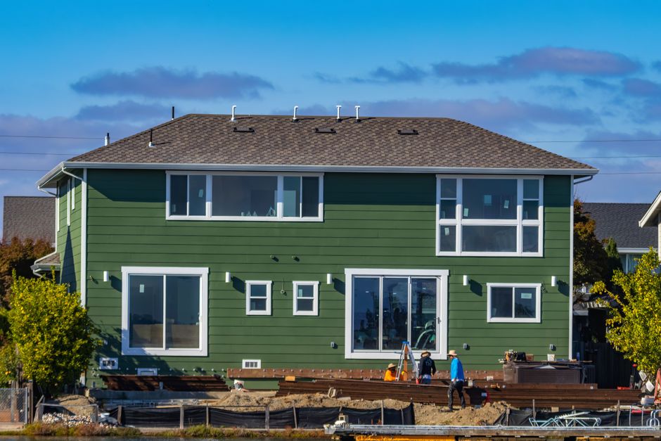 Green vinyl-sided house under construction with workers and materials visible